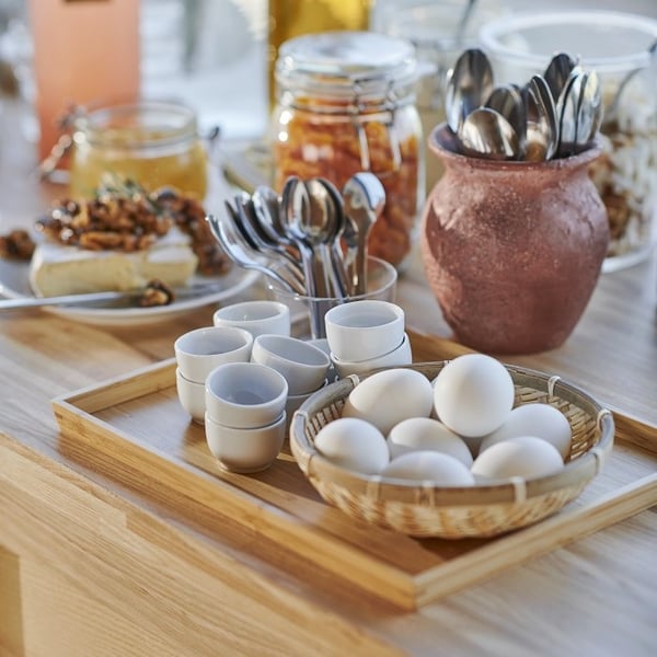 A close-up view of a breakfast buffet setup on a wooden surface. A wicker basket holds white hard-boiled eggs, placed on a wooden tray alongside several white ceramic egg cups. Next to them are glass containers filled with silver teaspoons, and a rustic terracotta pot also holding cutlery. In the background, a glass jar contains preserved fruit or vegetables, and a dessert plate with banana slices topped with nuts and dried fruit adds a sweet touch. The scene is warmly lit by natural daylight, creating a cozy, inviting atmosphere.