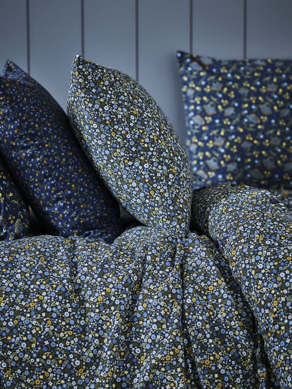 A close-up view of several decorative pillows and bedding with a floral pattern. The pillows are in shades of blue, yellow, and white, and they’re piled against a gray background.