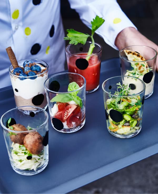 A closeup of a person holding a tray with 6 clear glasses filled with different courses of food.