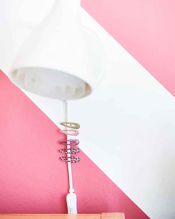 A closeup of a white reading lamp mounted to the wall, whose wire has a few hairclips attached to it for easy reach.