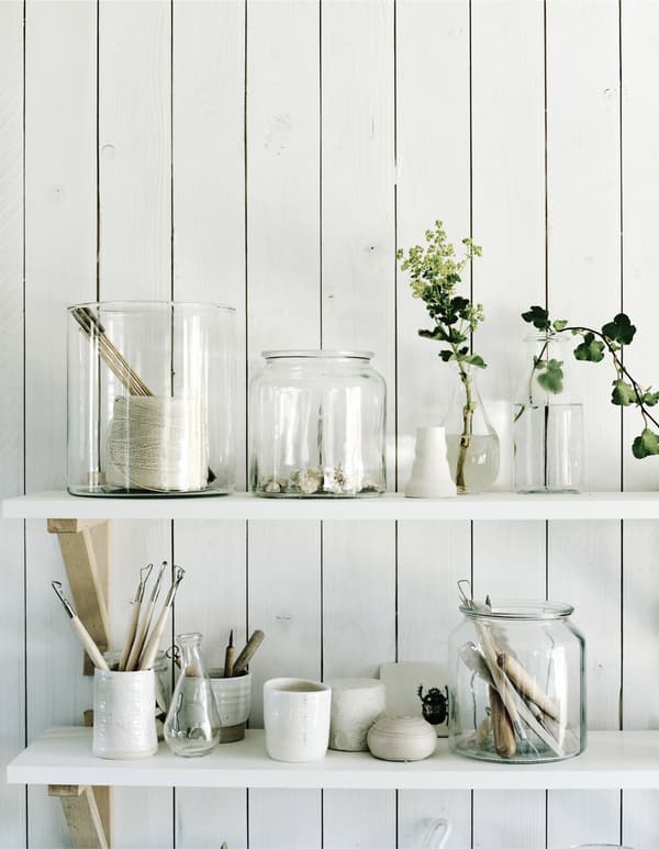 A closeup of two white shelves mounted to a white wooden wall, holding clear jars in Annika’s pottery studio.