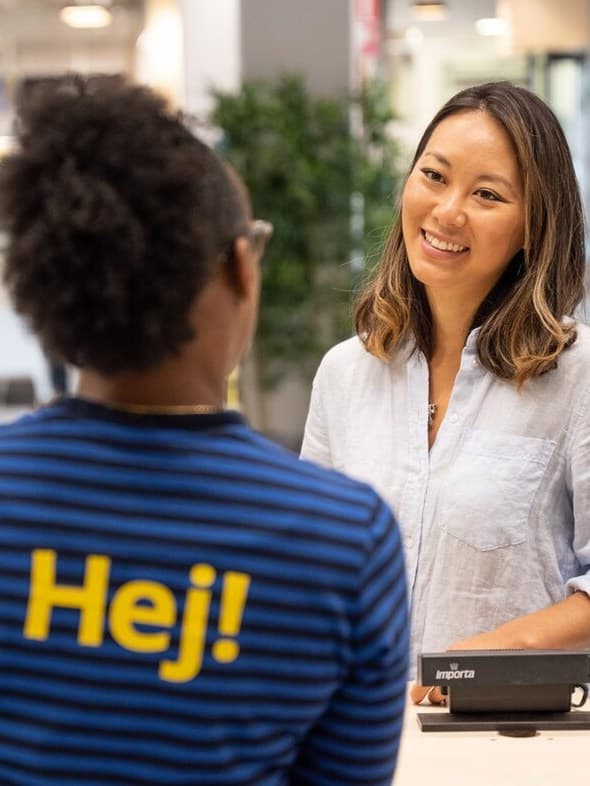 A co-worker in a blue top with the word "Hej!" speaks to a customer. The customer smiles at the co-worker.
