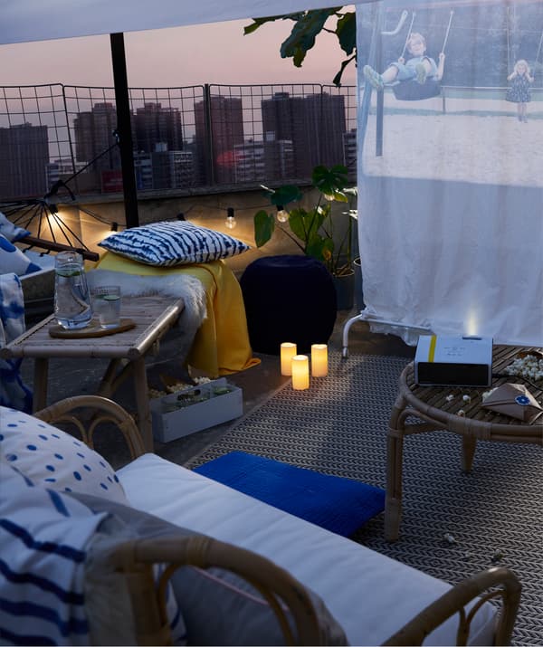 A coffee table with popcorn grouped with pouffes, outdoor armchairs and a hammock in front of a film screen on a rooftop.