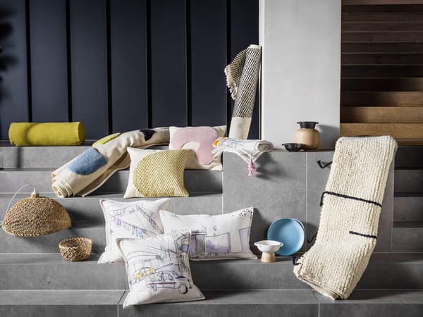 A collection of LOKALT products including pillows with an illustrated city, rugs, and a weaved lamp shade.