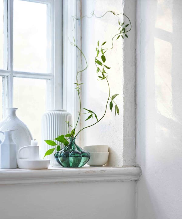 A collection of white ceramic pots sit on a windowsill with a green glass vase at their centre.