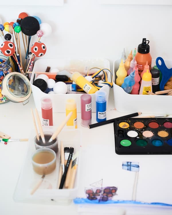 A collection of white plastic boxes organising paints, brushes and other children's arts and crafts supplies at a desk.