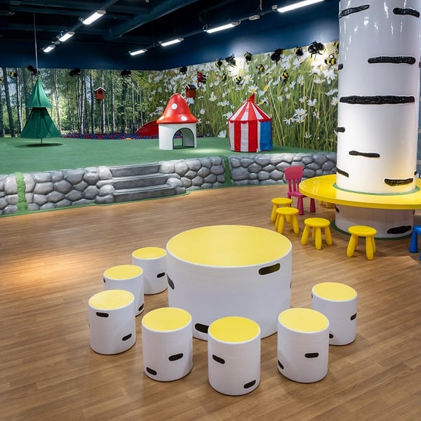 A colorful indoor children's play area featuring yellow-and-white tables with matching stools, a white tree-like column with a circular play table, and a raised grassy section with playful structures like a red mushroom house, a circus tent, and hanging green trees, set against a forest-themed wall mural.