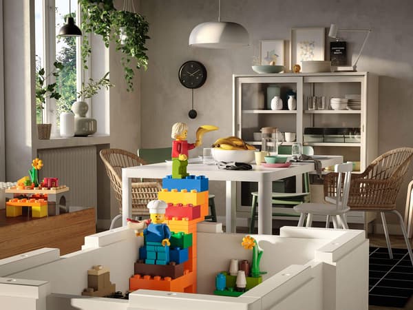 A colorful LEGO brick tower with two LEGO characters inside an open white storage box inside a dining room.
