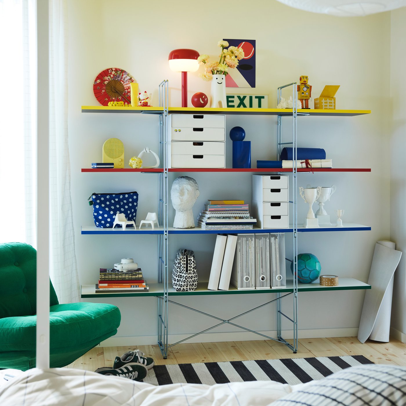 A colorful shelving unit with red, yellow, blue, and green shelves, filled with books, storage boxes, and decorative items in a modern, cozy room. A green chair and a striped rug are visible in the foreground, creating a playful and creative atmosphere.