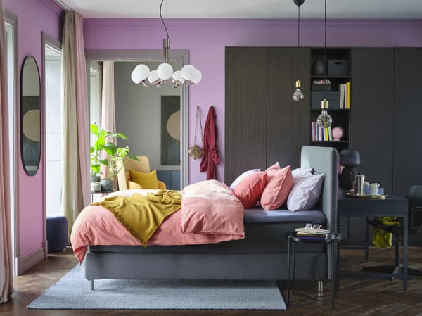 A colourful bedroom with lilac walls, light brown-red bed linen, a yellow throw, a black-brown wardrobe and a grey divan bed.