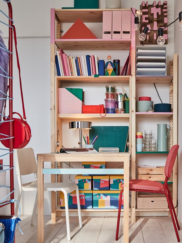 A colourful bedroom work/dining area with a foldable IKEA IVAR table in pine that's paired with red and white chairs.