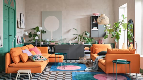 A colourful living room with a sofa and a chaise longue in orange, bold print rugs and green tray tables.