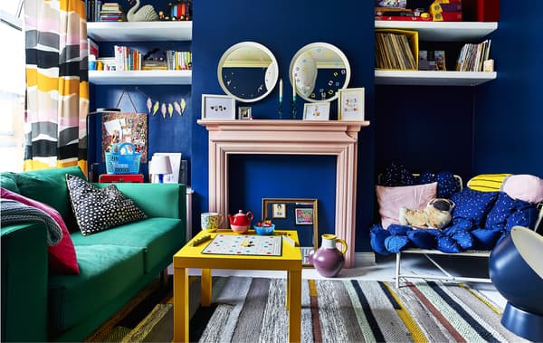 A colourful living room with green sofa and yellow coffee table in front of a pink fireplace mantel against navy blue walls.