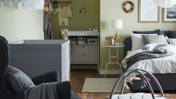 A combined bedroom and baby room with a gray upholstered bed, grey crib, grey changing table/chest of drawers, green walls.