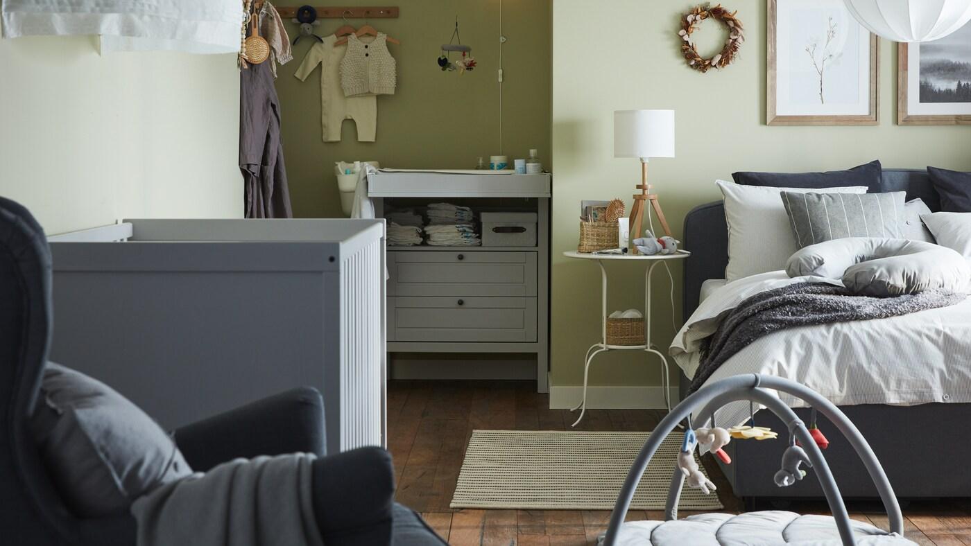 A combined bedroom and baby room with a grey upholstered bed, grey crib, grey changing table/chest of drawers, green walls.