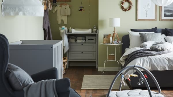 A combined bedroom and baby room with a grey upholstered bed, grey cot, grey changing table/chest of drawers, green walls.