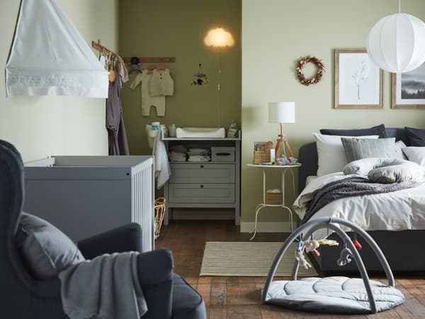 A combined bedroom and baby room with a grey upholstered bed, grey cot, grey changing table/chest of drawers, green walls.