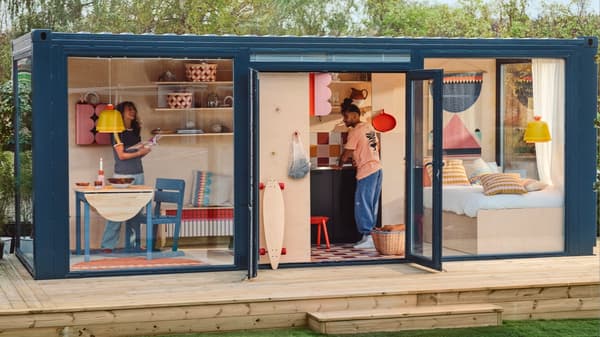 A compact living space showcasing OMMJÄNGE. The space shows large windows, a woman at a table and a man in the kitchen, with living, dining, and bedroom.