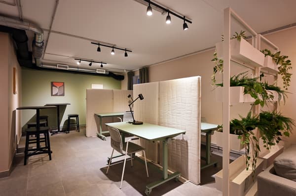 A contemporary co-working space with shared desks separated by acoustic divider panels, adjustable desk lamps, and shelving with indoor plants. Soft lighting, warm neutral tones, and industrial-style details create a calm and functional environment for focused work and collaboration.