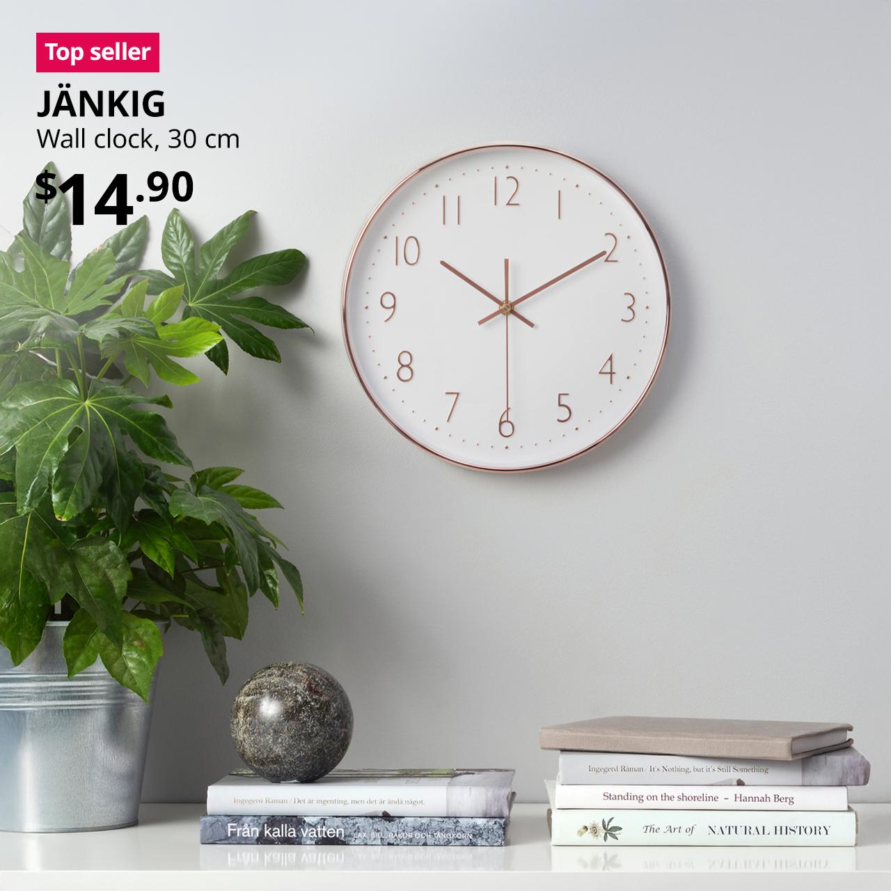 A copper coloured JÄNKIG wall clock hung above a wall shelves with decorations of plant pot and books. A red "Top seller" label is on the top left of image, with black text below that reads: JÄNKIG, Wall clock, 30 cm. $14.90