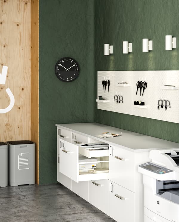 A copy room with plenty of white drawers, white pegboards, white spotlights, waste sorting bins and a black wall clock.
