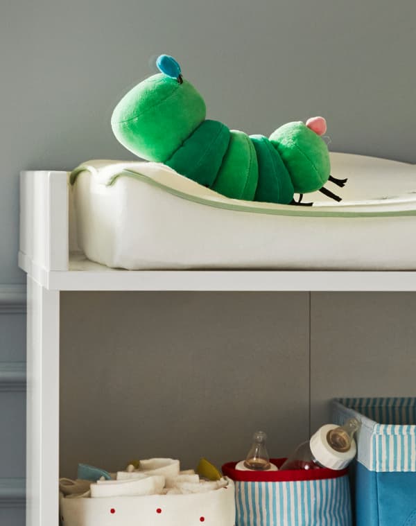 A corner of a baby changing mat with KLAPPA caterpillar soft toy on it and baby milk bottles and cloths in baskets on the shelf below.