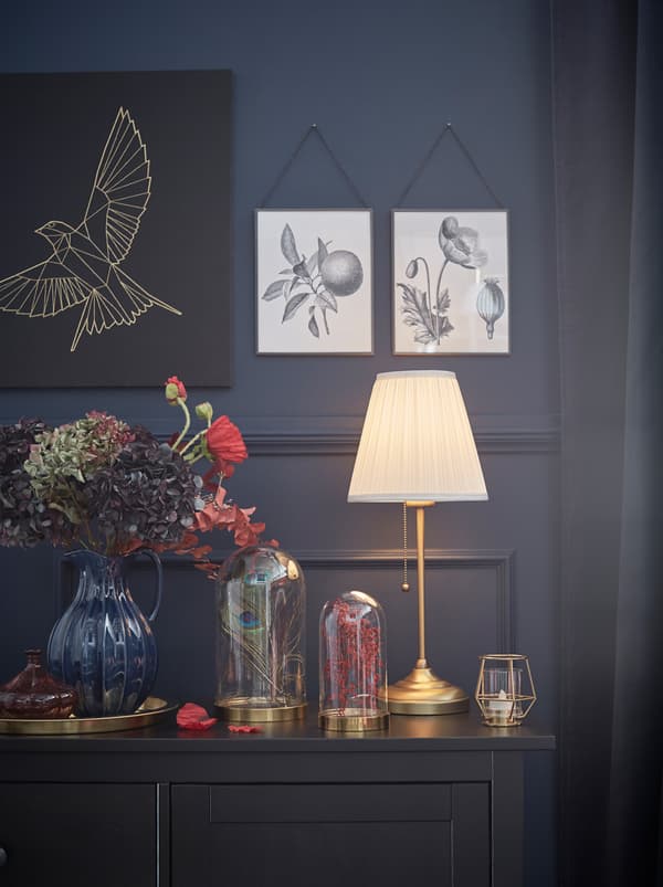 A corner of a dark-panelled room with art and decorations on both walls and surfaces, all softly lit by an ÅRSTID lamp.
