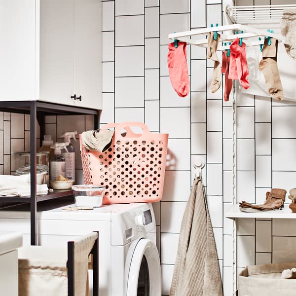 A corner of a laundry room with white hanging drying rack, washing machine, pink SLIPP flexible basket and white tiles.