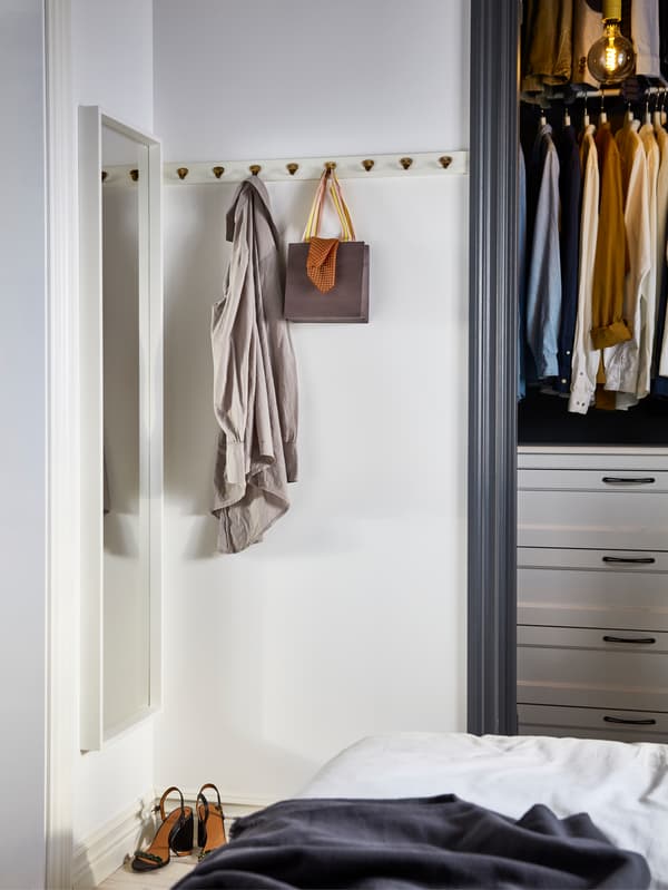 A corner of a white bedroom with a vertically hung IKEA NISSEDAL white mirror and a row of KVASP brass-coloured hooks.
