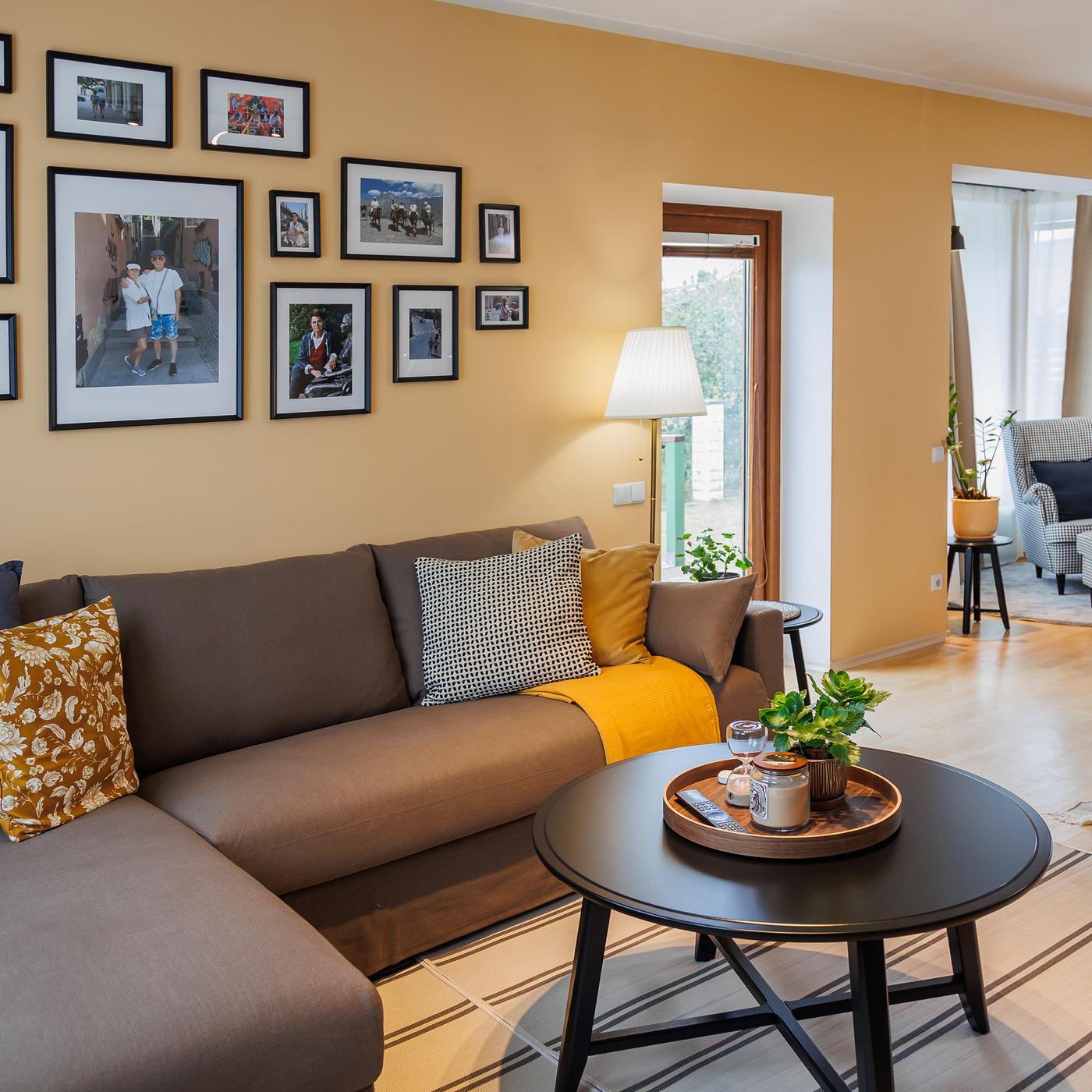 A corner of the living room features a brown three-seat sofa with patterned cushions and a round black coffee table. On the wall above the sofa hangs a collage of framed pictures.