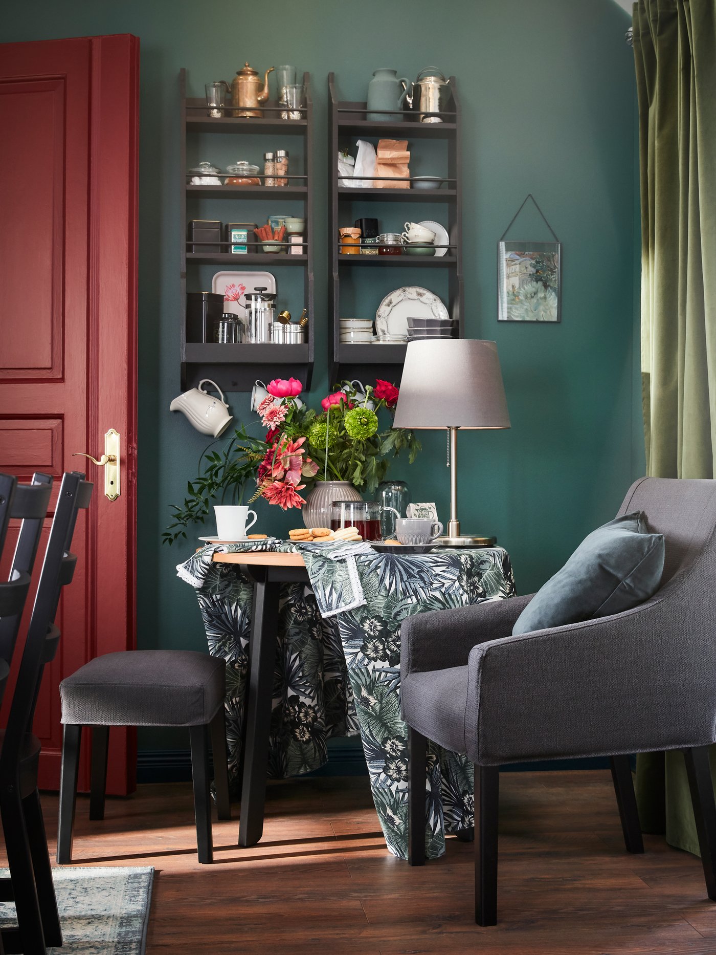 A corner with a black/dark gray chair with armrests, a table where tea is served and a window with white/green curtains.