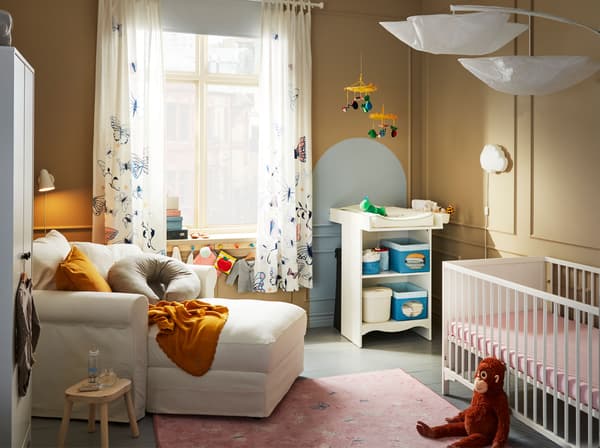 A cosy baby room, with a white IKEA GRÖNLID chaise longue, an IKEA SOLGUL changing table, a white cot, soft toys and mobiles.