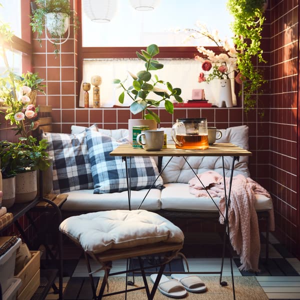 A cosy balcony features a black TÄRNÖ outdoor table, surrounded by plants, cushions, and a glass pot with tea.