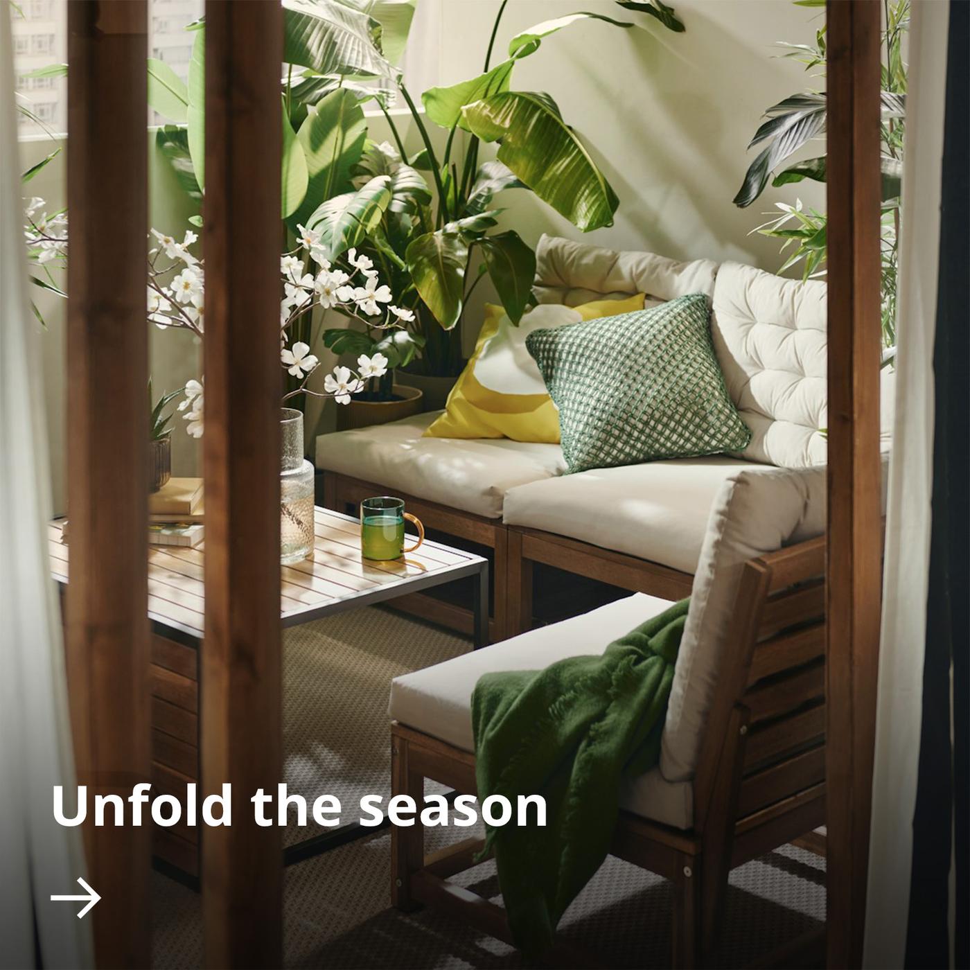 A cosy balcony seating area with wooden furniture, beige cushions, and green accents, surrounded by lush potted plants in warm sunlight.