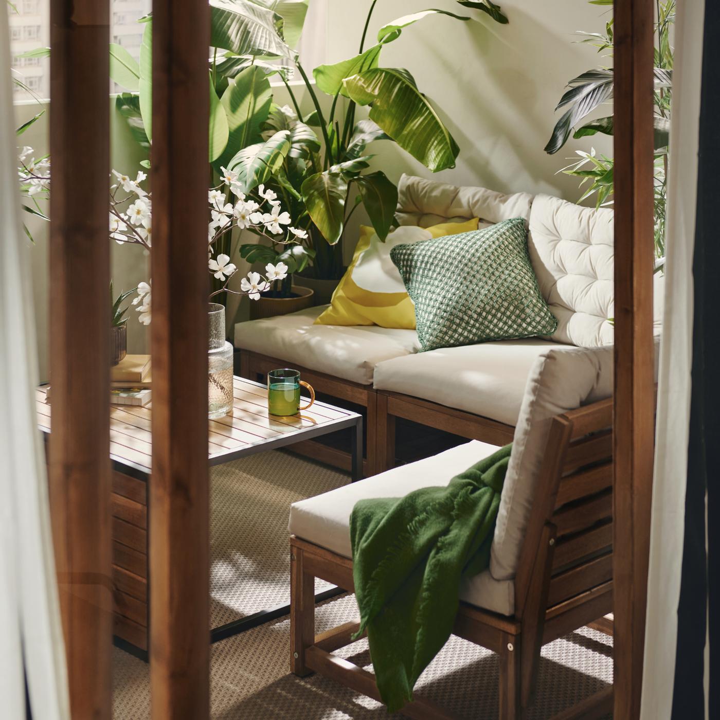 A cosy balcony seating area with wooden furniture, beige cushions, and green accents, surrounded by lush potted plants in warm sunlight.