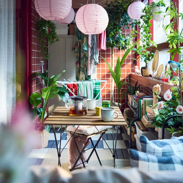 A cosy balcony with green plants and pink lamps, featuring a black and light brown wooden TÄRNÖ outdoor table and tea mugs.