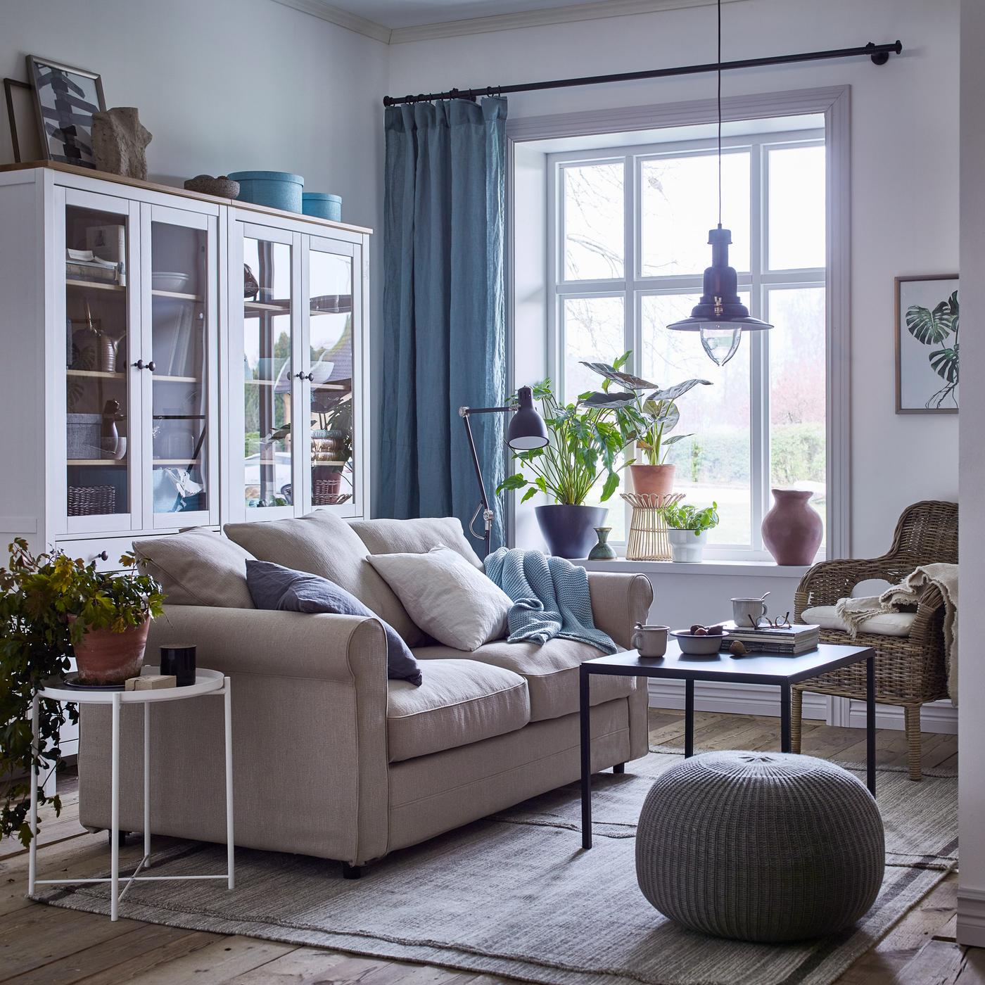 A cosy beige and white living room with blue curtains and a GRÖNLID 2-seat sofa in Sporda natural beside a rattan chair and glass storage cabinets.
