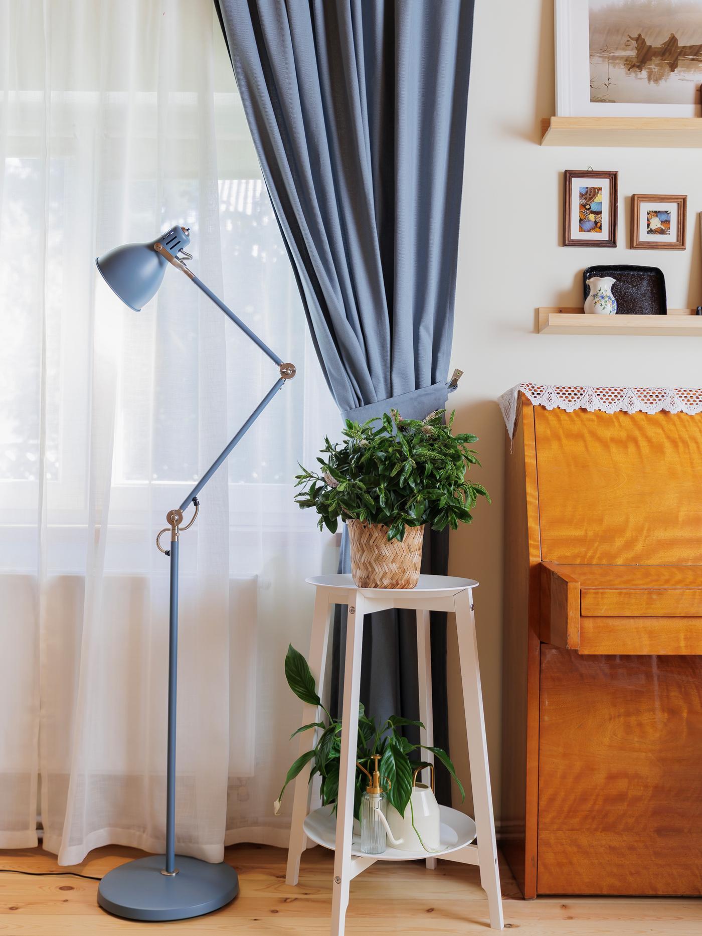 A cosy corner with a wooden piano by a window dressed with sheer white curtains and blue drapes, beside a tall floor lamp and a white plant stand holding a green plant.