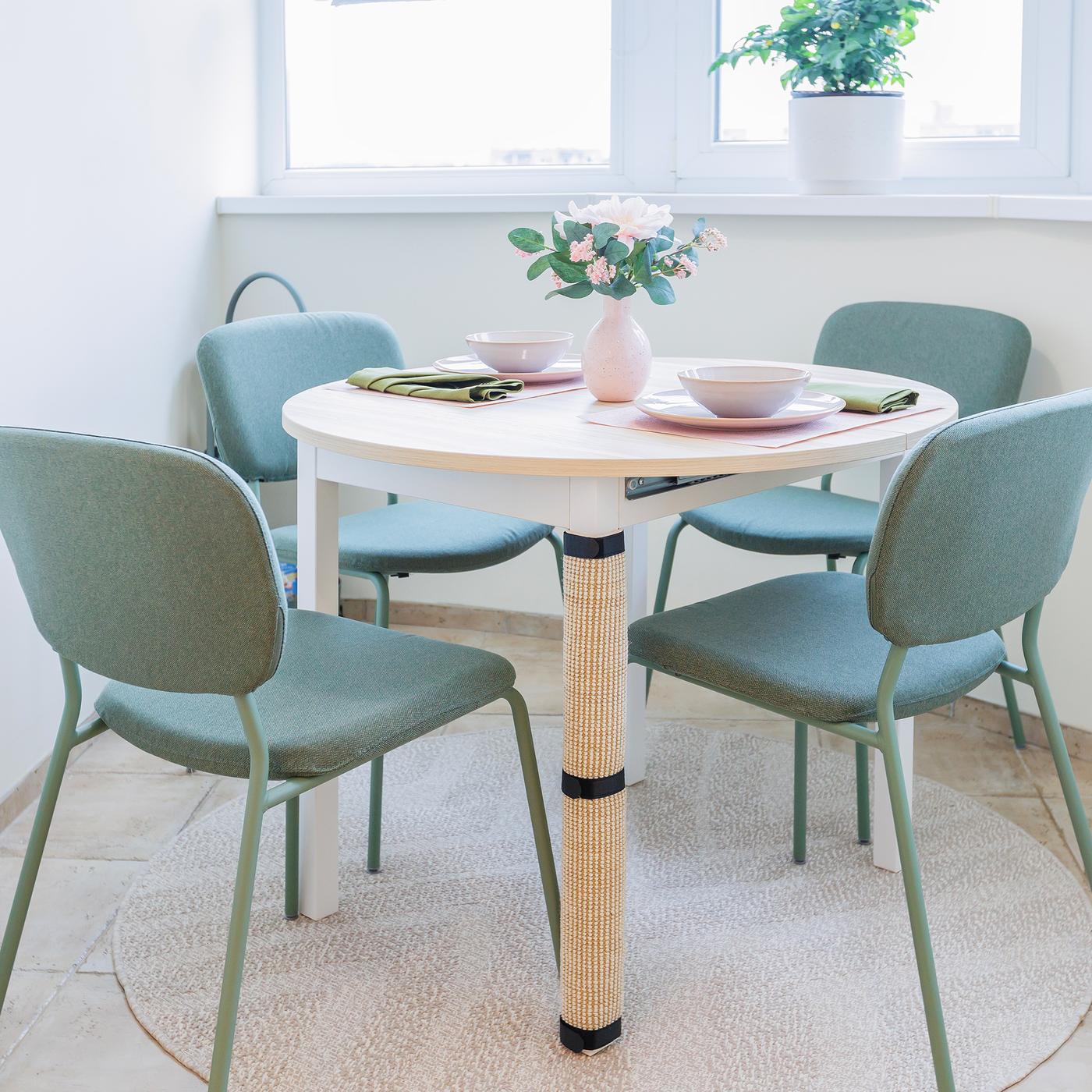 A cosy dining nook with a round wooden table and four green chairs, placed by a bright window with potted plants on the sill.