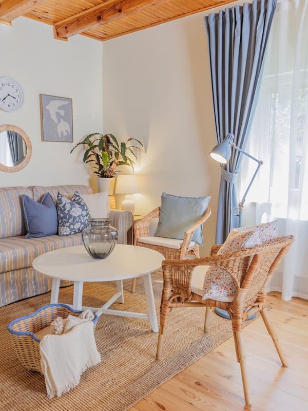 A cosy living room corner featuring a white coffee table with a woven basket beneath, surrounded by rattan armchairs, a striped sofa-bed and a floor lamp.
