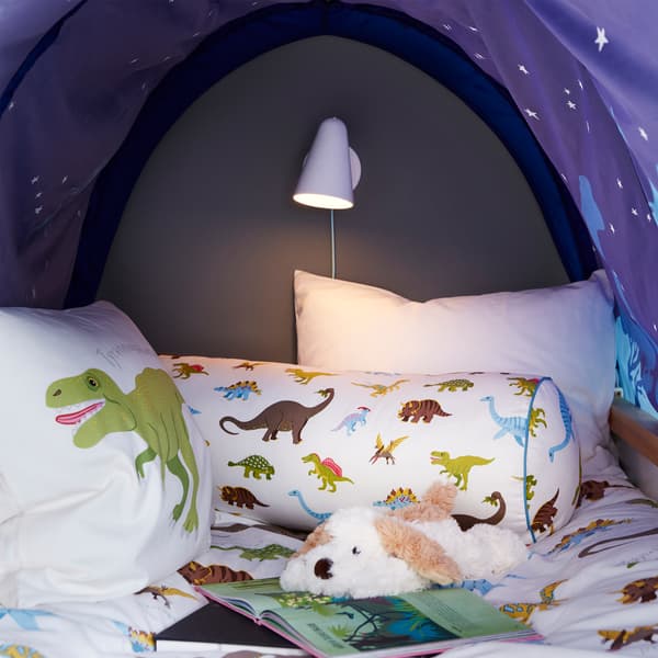 A cosy nook with a blue bed tent, a dog soft toy, a white wall lamp, an open book and cushions with dinosaur patterns.