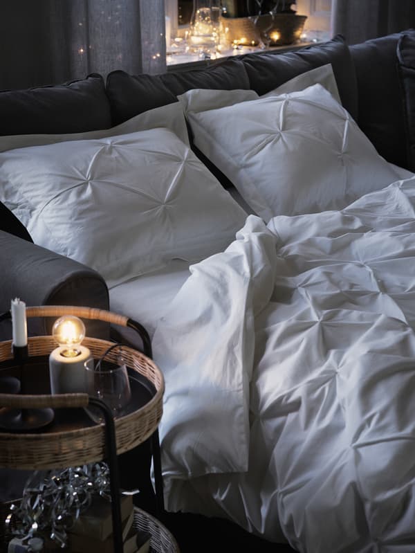 A cosy white double bed is made up in a grey GRÖNLID sofa-bed, with a wicker LUBBAN trolley beside it.