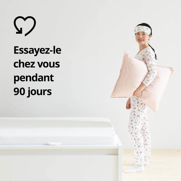 À côté d’un lit, femme en pyjama tenant un oreiller. Sur l’image, icône en forme de cœur et le texte « Essayez-le à la maison pendant 90 jours ».