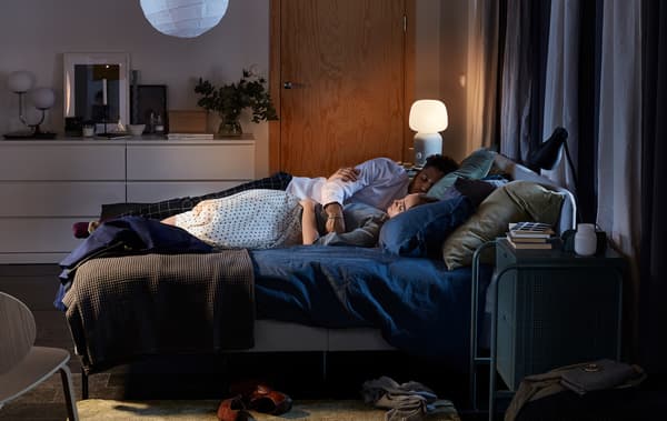 A couple cuddle asleep in a SLATTUM upholstered bed, with a SYMFONISK lamp/speaker lit in the background.