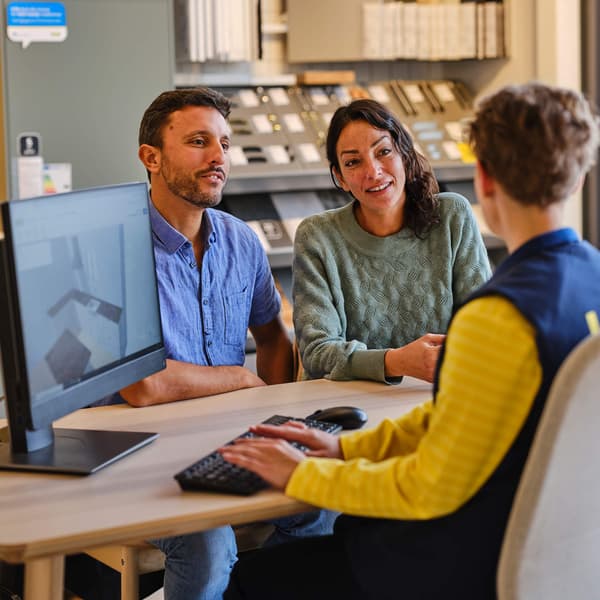 A couple sits at a desk with a store employee, viewing a computer screen and discussing home design options in a showroom.