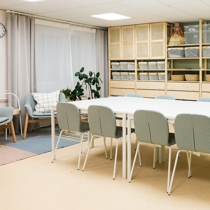 A cozy and functional room for holding workshops with a long table and gray-green chairs around it. Wooden shelves with matching boxes and baskets for storing materials on the wall.