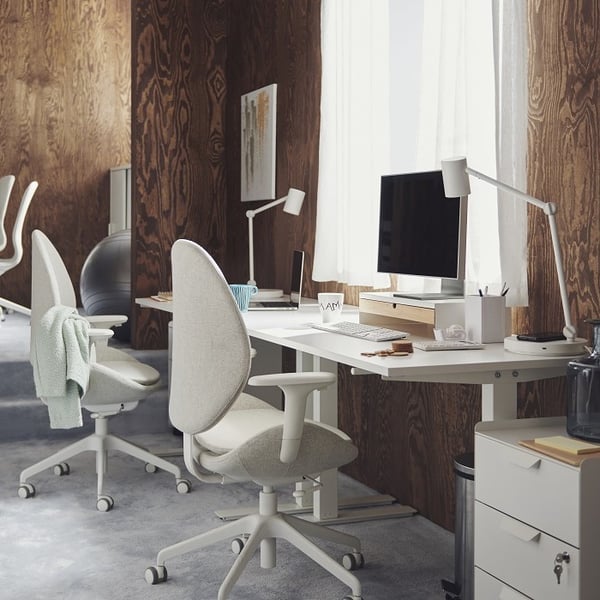 A cozy and modern home office setup with a dual workspace arrangement. Two ergonomic office chairs with high curved backs and soft upholstery face a long white desk positioned against a dark wood-paneled wall. The desk is equipped with minimalist white desk lamps, a computer monitor on a wooden riser, stationery, books, and a ceramic cup labeled 