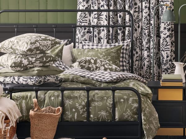 A cozy bedroom with a black metal bed frame, green patterned bedding, and matching curtains. Decorative pillows and woven baskets add to the homey ambiance.
