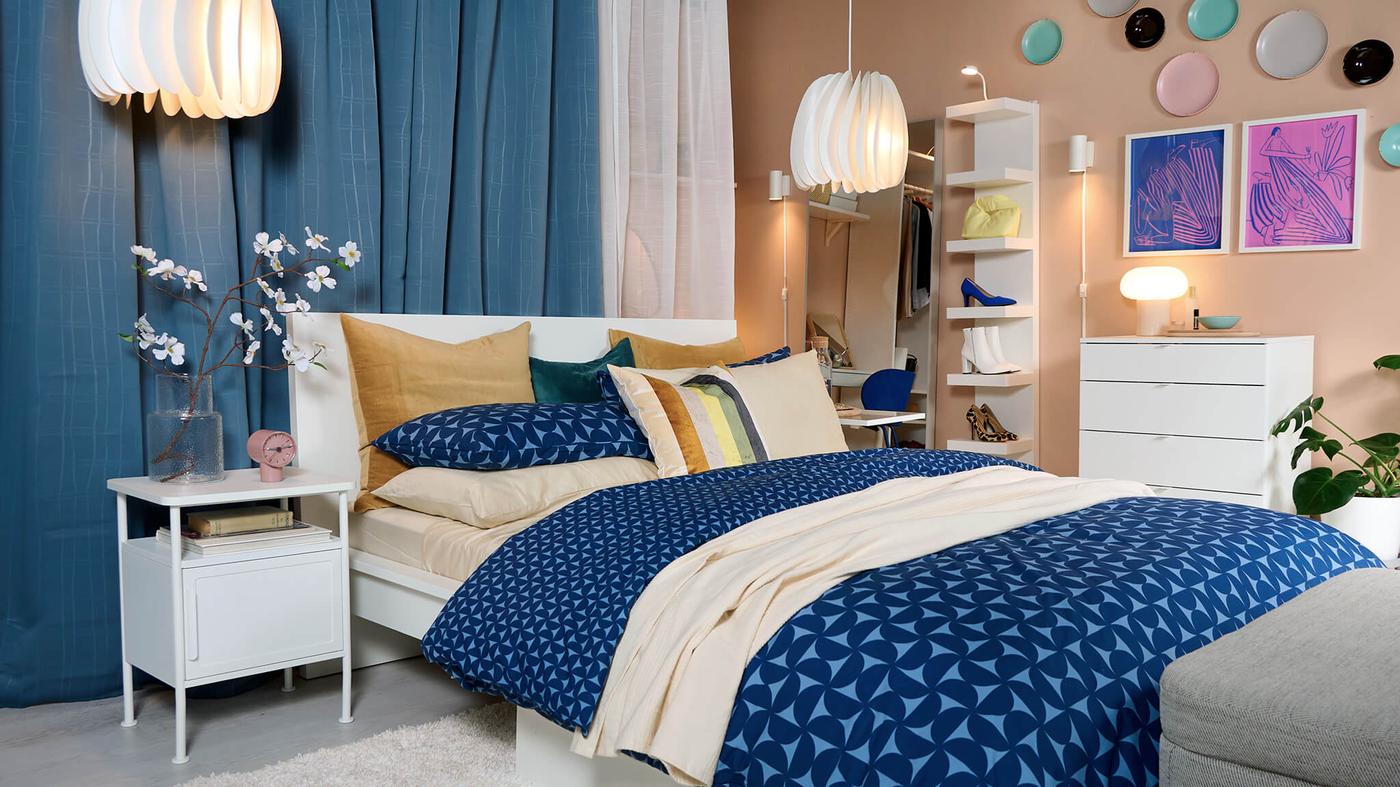 A cozy bedroom with blue and beige bedding, modern decor, pendant lights, and a white dresser against a peach-toned wall.