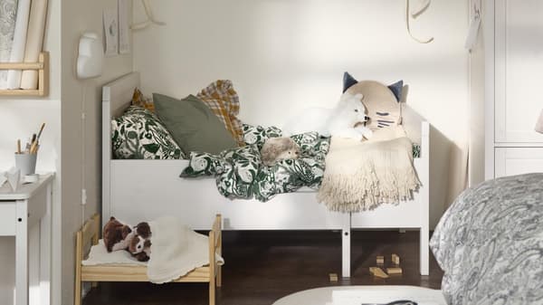 A cozy children's room with a small bed, green and white bedding, stuffed animals, and a miniature wooden bed for toys on a dark wooden floor.